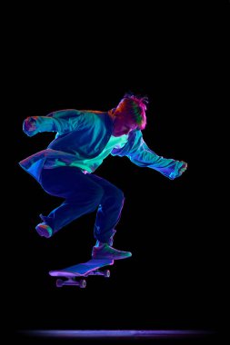 Mavi kotlu tişörtlü ve şapkalı genç çocuk kaykayla antrenman yapıyor, neon ışıkta siyah stüdyo geçmişine karşı dublörlük yapıyor. Profesyonel spor kavramı, rekabet, eğitim, eylem.
