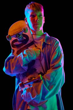 Gündelik giysiler içinde kaykaylı genç bir çocuk neon ışıkta siyah stüdyo geçmişine karşı poz veriyor. Profesyonel spor kavramı, rekabet, eğitim, eylem. İlan için alanı kopyala