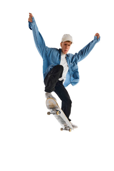 Teen boy in blue jeans shirt and cap in motion, training with skateboard, doing stunts isolated over white background. Concept of professional sport, competition, training, action. Copy space for ad