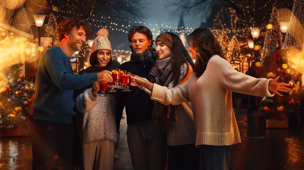 Cozy winter fair with lights. Young happy people, friends meeting, drinking mulled wine, celebrating holidays outdoors. Concept of winter holidays, Christmas, traditions, outdoor fair, happiness