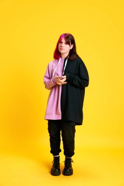 Alışılmadık boyalı saçlı, iki renkli, açık renk makyajlı ve canlı sarı stüdyo geçmişine karşı sakin yüzüyle ayakta duran genç bir kız. Gençlik kavramı, kendini ifade etme, moda, duygular