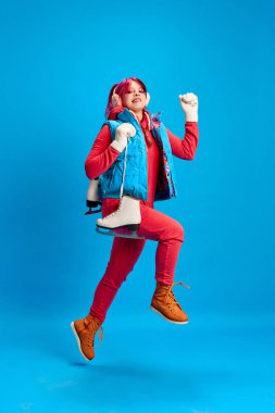 Sıcak kış kıyafetleri içindeki neşeli genç kız mavi stüdyo arka planında paten delikleri açıyor. Aktif hafta sonları. Gençlik kavramı, kendini ifade etme, kış tatili, aktif yaşam tarzı, duygular