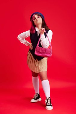 Parlak makyajlı, piercingli, gözlüklü, şık elbiseli ve bereli genç bir kız kırmızı stüdyo arka planında poz veriyor. Gençlik kültürü, kendini ifade etme, eğitim, moda, duygu kavramı