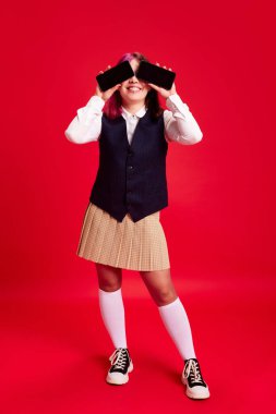 Genç bir kız, üniformalı bir öğrenci kırmızı stüdyo geçmişine karşı iki cep telefonuyla gözlerini kapatıyor. Gençlik kültürü, kendini ifade etme, eğitim, moda, duygu kavramı