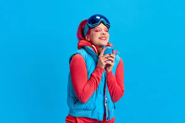Warming up with hot tea. Smiling young girl with bright makeup wearing winter warm clothes over blue studio background. Concept of youth, self-expression, winter vacation, active lifestyle, emotions