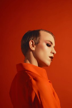 Şık saç stili, yazı makyajı, ruj ve kirpikleriyle kırmızı stüdyo arka planına poz veren müsrif bir genç. Erkek makyaj anlayışı, moda, lgbtq topluluğu, kişisel kimlik, kabullenme