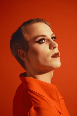 Şık saç stili, yazı makyajı, ruj ve kirpikleriyle kırmızı stüdyo arka planına poz veren müsrif bir genç. Erkek makyaj anlayışı, moda, lgbtq topluluğu, kişisel kimlik, kabullenme