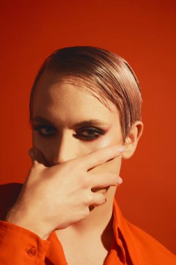 İbne. Kırmızı stüdyo arka planında parlak makyajlı ve saç stili olan genç adamın portresi. Kozmetik. Erkek makyaj anlayışı, moda, lgbtq topluluğu, kişisel kimlik, kabullenme