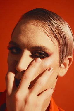 İbne. Kırmızı stüdyo arka planında parlak makyajlı ve saç stili olan genç adamın portresi. Kozmetik. Erkek makyaj anlayışı, moda, lgbtq topluluğu, kişisel kimlik, kabullenme