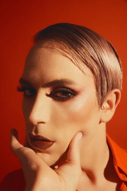 Renkli ceketli ve parlak makyajlı genç transseksüel bir adamın portresi kırmızı stüdyo arka planında poz veriyor. Erkek makyaj anlayışı, moda, lgbtq topluluğu, kendini tanıtma