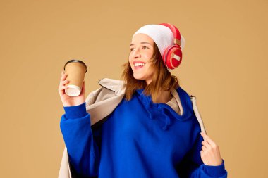 Gülümseyen, şık giyinen, kulaklıkla müzik dinleyen ve bej arka plana karşı gelmek için kahve içen genç bir kız. Gençlik kültürü, moda, insani duygular, yaşam tarzı, müzik kavramı