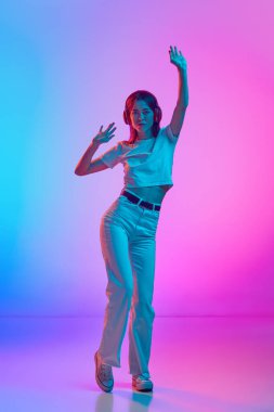 Gündelik giysiler içinde, kulaklıkla müzik dinleyen ve neon ışıklarıyla dans eden, gradyan pembe renkli stüdyo arka planında dans eden güzel bir kadın. İnsan duyguları, yaşam tarzı, gençlik kültürü kavramı