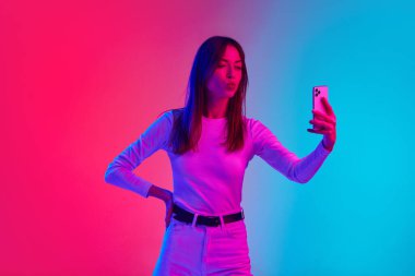 Genç ve güzel bir kadın cep telefonuyla selfie çekiyor. Neon ışıklarıyla çekilen pembe renkli stüdyo arkaplanına karşı. Sosyal medya. İnsan duyguları kavramı, yaşam tarzı, gençlik kültürü, yüz ifadesi