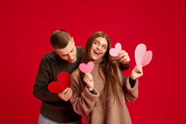 Genç mutlu adam ve kadın, çift, erkek arkadaş ve kız arkadaş kırmızı stüdyo geçmişine karşı kağıt kalplerle poz veriyorlar. Aşk kavramı, ilişki, Sevgililer Günü, duygular, yaşam tarzı