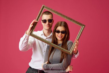 Genç bir çift, güneş gözlüklü bir adam ve bir kadın pembe stüdyo arka planında fotoğraf çerçevesiyle poz veriyorlar. Bağ kurmak. Aşk kavramı, ilişki, Sevgililer Günü, duygular, yaşam tarzı