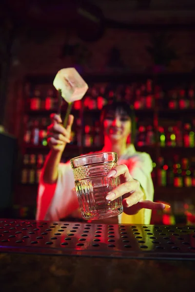 Young woman, bartender putting ice cubes into glass, making delicious cocktail in bar in neon light. Gin garden. Concept of occupation, nightlife, bar, party, alcohol drink, mixologist