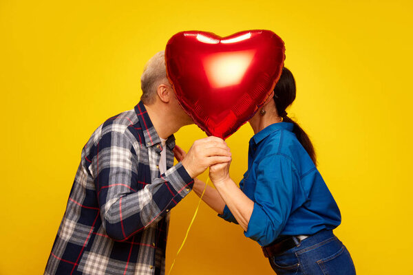 Старшая пара, жена и муж стоят позади красного шара в форме сердца и целуются на фоне жёлтой студии. О браке, отношениях, Дне святого Валентина, любви, эмоциях, моде