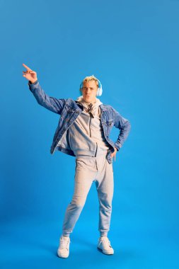 Kot ceketli, spor kıyafetli, kulaklıklı müzik dinleyen ve mavi stüdyo arka planında dans eden neşeli, şık bir genç. İnsan duyguları, gençlik, pozitiflik, gündelik moda kavramı