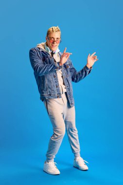 Kot ceketli, spor kıyafetli, kulaklıklı müzik dinleyen ve mavi stüdyo arka planında dans eden neşeli, şık bir genç. İnsan duyguları, gençlik, pozitiflik, gündelik moda kavramı