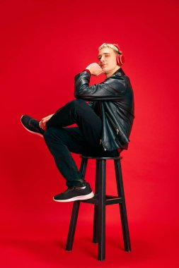 Deri ceketli, kot pantolonlu, kırmızı stüdyo arka planında kulaklıkla müzik dinleyen yakışıklı bir genç. İnsan duyguları, gençlik, moda, yaşam tarzı, pozitiflik kavramı
