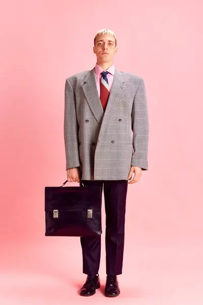 Portrait of young man in formal wear, jacket and glasses, standing with briefcase against pink studio background. Serious face. Concept of emotions, business, profession and occupation, lifestyle