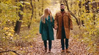 Güzel genç bir çift, kadın ve erkek el ele tutuşuyor, gülümsüyor, konuşuyor ve ılık bir günde sonbahar ormanında yürüyor. Sonbahar mevsimi kavramı, ilişki, hafta sonu aktivitesi, doğa keyfi