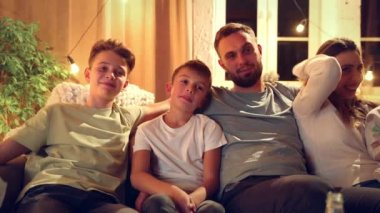 Gülümseyen ebeveynler ve çocuklar akşamları oturma odasında kanepede oturuyor ve birlikte televizyon izliyorlar. Komedi filmi ya da gösteri. Aile kavramı, boş zaman, rahatlama, çocukluk ve ebeveynlik