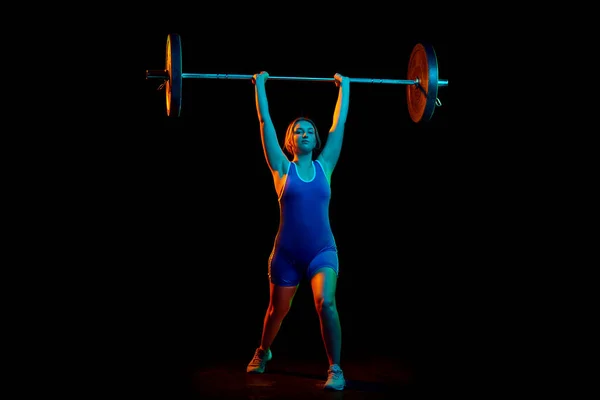 Full-length image of professional athlete, young woman training, lifting heavy weight, barbell against black background in neon light. Concept of sport, strength, healthy lifestyle, power, endurance