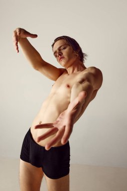 Kaslı, formda, spor tişörtsüz, gri stüdyo geçmişine karşı iç çamaşırlarıyla duran genç bir adam. Erkeklerin güzellik, sağlık, vücut bakımı, spor yaşam tarzı kavramı