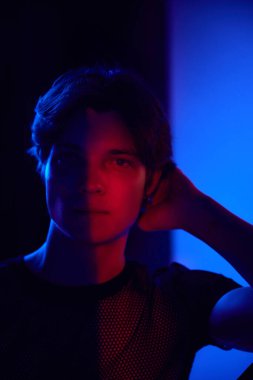 Kameraya bakan yakışıklı bir gencin portresi, neon ışıkta koyu mavi arka planda poz veriyor. Gölgeler. Gençlik kültürü, moda ve erkek güzelliği, duygular, ilham, eğilimler kavramı