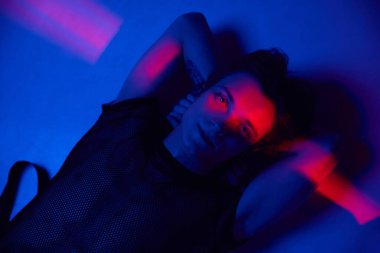 Yakışıklı genç bir adamın portresi, erkek model yerde yatıyor ve koyu mavi arka planda neon ışıkla poz veriyor. Gençlik kültürü, moda ve erkek güzelliği, duygular, ilham, eğilimler kavramı