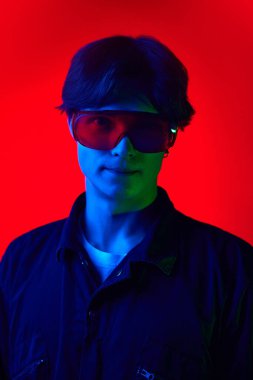 Koruyucu gözlüklü yakışıklı genç adamın portresi parlak kırmızı arka planda mavi ışıkta poz veriyor. Gençlik kültürü, moda ve erkek güzelliği, duygular, ilham, eğilimler kavramı