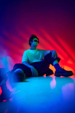 Şık giyinen, beyaz tişörtlü, güneş gözlüklü, neon ışıklı, renkli arka planda poz veren şık bir genç. Gençlik kültürü, moda ve erkek güzelliği, duygular, ilham, eğilimler kavramı