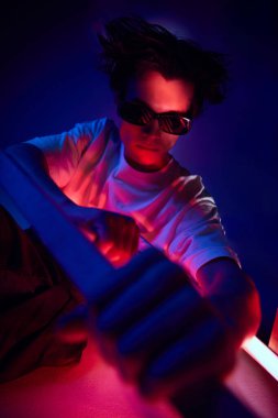 Fantezi, siber punk. Günlük kıyafetler ve güneş gözlüğü takan yakışıklı genç adam mavi arka planda neon ışıkta poz veriyor. Gençlik kültürü, moda ve erkek güzelliği, duygular, ilham, eğilimler kavramı