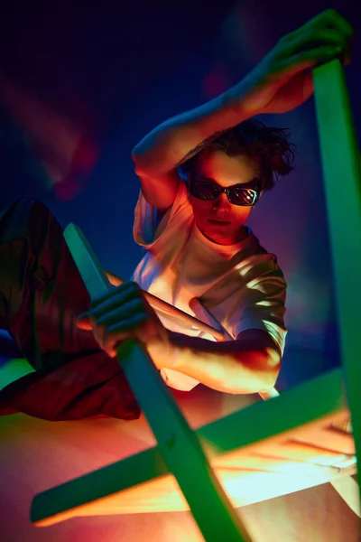 Fantasy, cyberpunk. Handsome young man in casual clothes and sunglasses posing over blue background in neon light. Concept of youth culture, fashion and male beauty, emotions, inspiration, trends