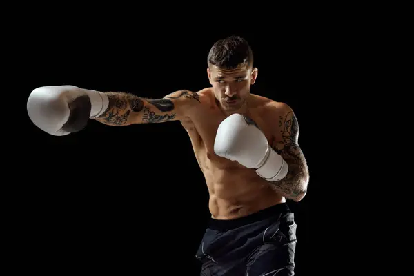 Concentrated serious young man, shirtless boxing athlete with strong muscular body fighting isolated over black background. Concept of professional sport, combat sport, martial arts, strength