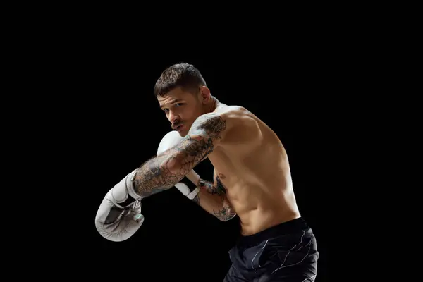 Strong, muscular young man, boxing athlete in gloves, training, fighting isolated over black background. Concept of professional sport, combat sport, martial arts, strength
