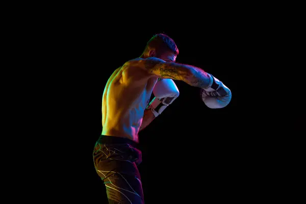 Young muscular man, boxing athlete in motion, training, fighting on ring isolated over black background in neon light. Concept of professional sport, combat sport, martial arts, strength