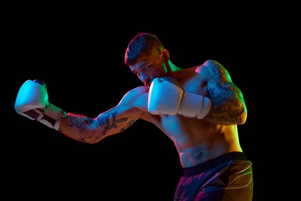Strong, muscular young man, boxing athlete in gloves, training, fighting isolated over black background in neon light. Concept of professional sport, combat sport, martial arts, strength