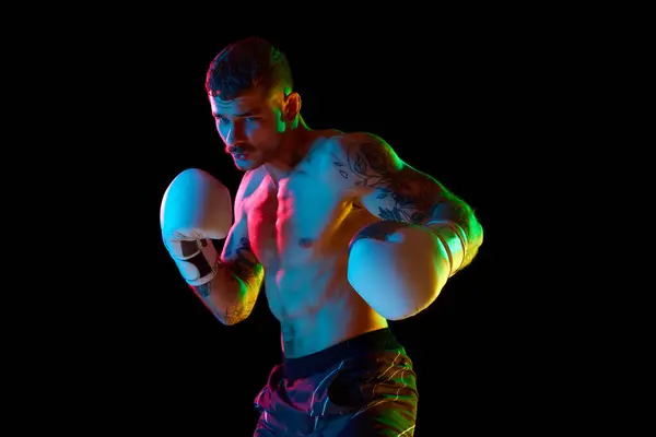Competitive, concentrated strong, muscular young man, boxing athlete training shirtless isolated on black background in neon light. Concept of professional sport, combat sport, martial arts, strength