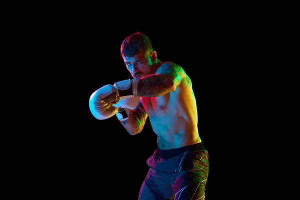 Young man, shirtless boxing athlete with muscular body training, punching isolated over black background in neon light. Concept of professional sport, combat sport, martial arts, strength