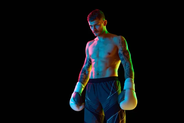 Young man with muscular strong shirtless body, boxing athlete standing isolated over black background in neon light. Concept of professional sport, combat sport, martial arts, strength