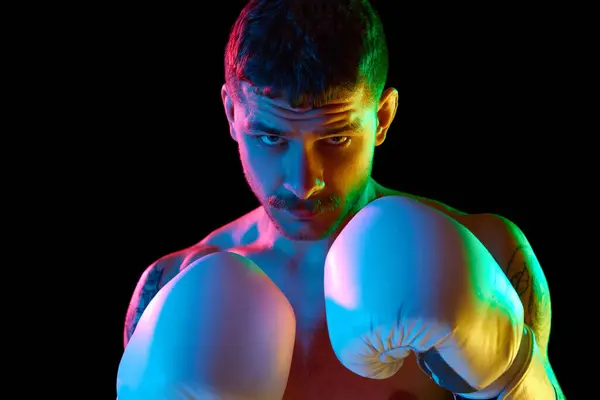 Portrait of concentrated, serious, brutal young man, boxing athlete standing shirtless with gloves isolated over black background in neon light. Concept of sport, combat sport, martial arts, strength