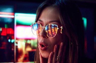 Mutlu ve heyecanlı genç kadın vitrinlere bakıyor gözlüklerinde neon ışıkları var. Alışveriş koliği, satış. İnsan duyguları, alışveriş, satış, tatil kavramı