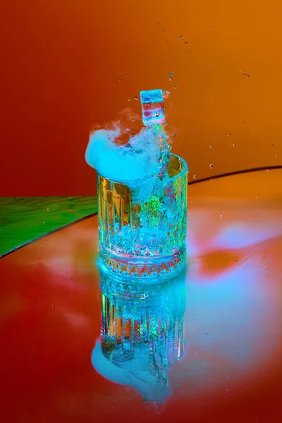 Ice cube falling down into glass with alcohol against orange background in neon light with smoke. Refreshment. Concept of alcohol drink, nightclub, party, taste, celebration.