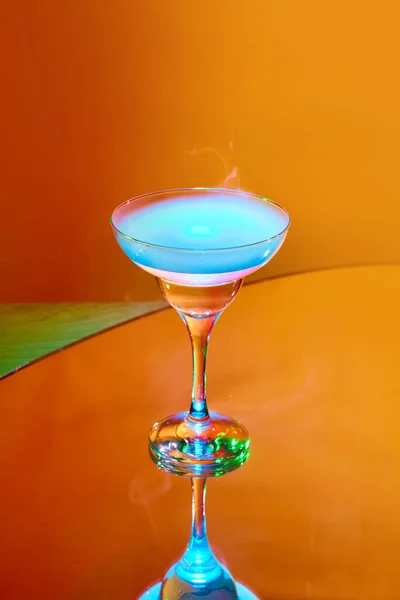 Extravagant drink serving. Glass with martini cocktail and smoke on top against orange background in neon light. Concept of alcohol drink, nightclub, party, taste, celebration.
