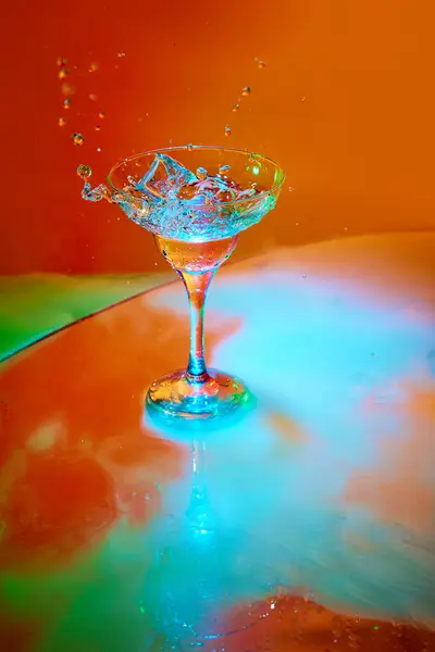 Ice cube falling into glass with martini cocktail against orange background in neon light with smoke effect. Splashes of drink. Concept of alcohol drink, nightclub, party, taste, celebration.