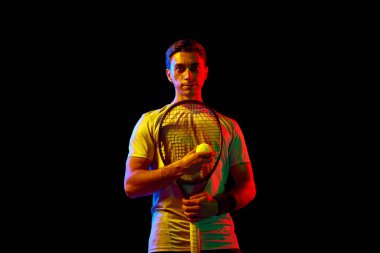 Atletik adam portresi, ayakta duran tenisçi, raket ve topla neon ışıkta karanlık arka planda poz veriyor. Profesyonel spor, rekabet, oyun, matematik, hobi, eylem kavramı