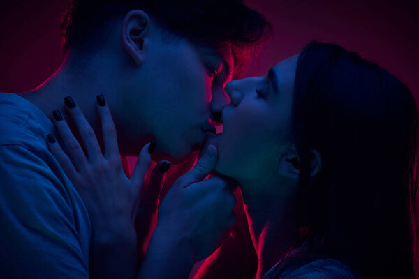 Portrait of passionate young man and woman, couple kissing, expressing love against purple background in neon light. Concept of romance, love, relationship, passion, youth, dating, happiness