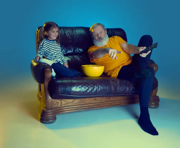 Happy senior man, grandfather sitting on couch at home with his little granddaughter and watching TV over blue background in neon light. Concept of family, happiness, care and love, emotions, leisure
